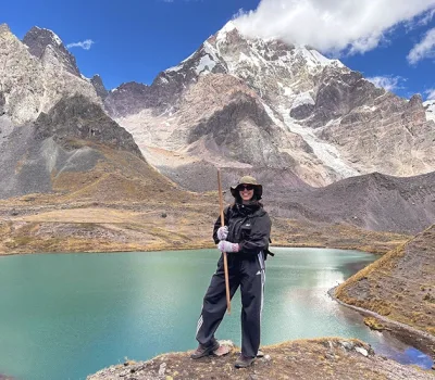 Female traveler posing with trekking gear by the turquoise waters of Pucacocha Lake beneath the snow-capped Ausangate Mountain in Cusco, Peru