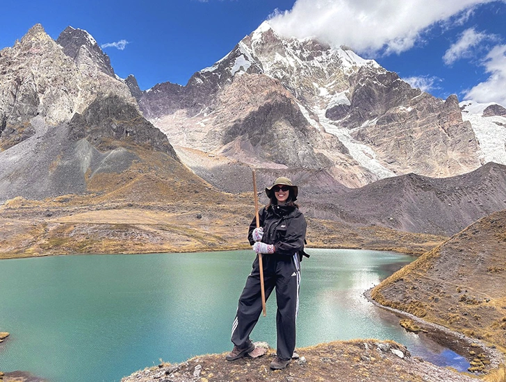Female traveler posing with trekking gear by the turquoise waters of Pucacocha Lake beneath the snow-capped Ausangate Mountain in Cusco, Peru